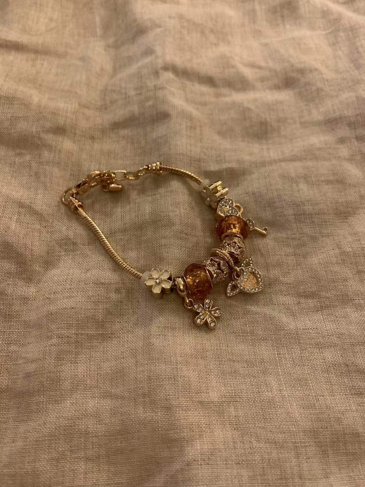 Gold charm bracelet with floral and heart charms, amber beads, and crystal accents on textured fabric