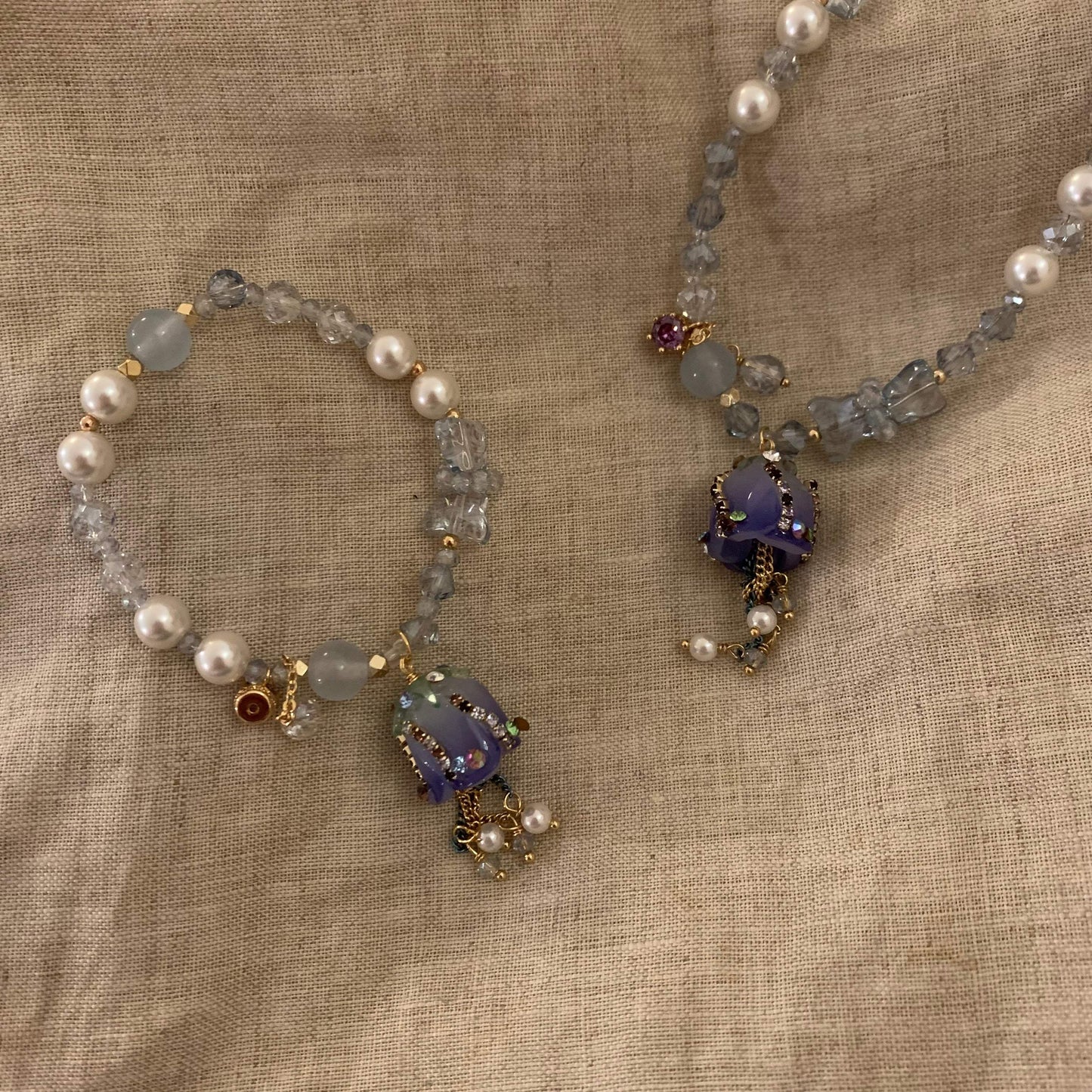 Handcrafted pearl and crystal necklace with purple pendant, gold accents, and delicate charms on linen background