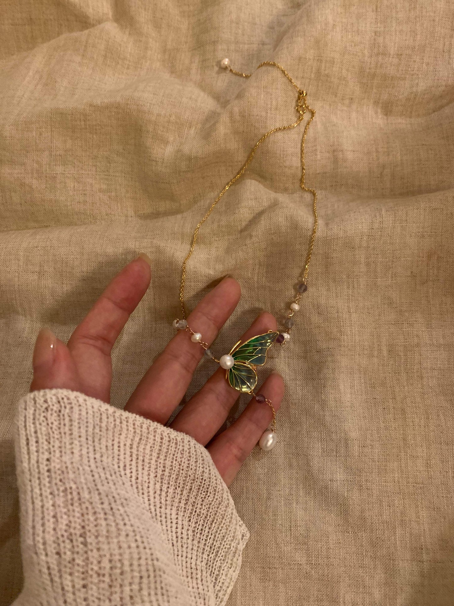 Gold butterfly necklace with green enamel wings, pearls, and gemstones on a delicate chain, held over linen fabric.