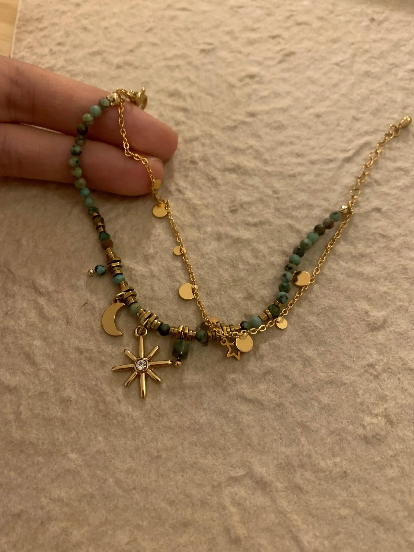 Gold celestial necklace with green beads, moon, star, and sunburst charms on textured fabric