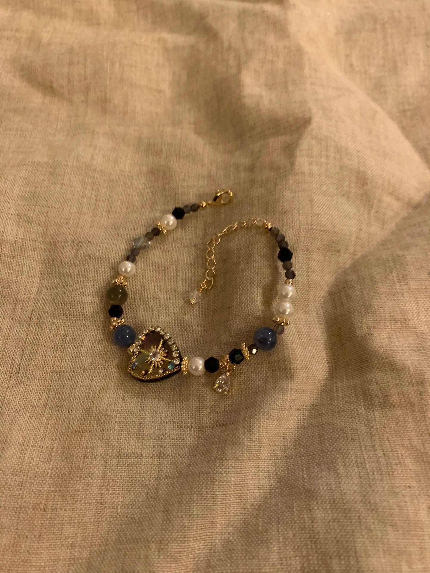 Elegant beaded bracelet with gold chain, pearls, and a heart-shaped pendant, displayed on linen fabric.