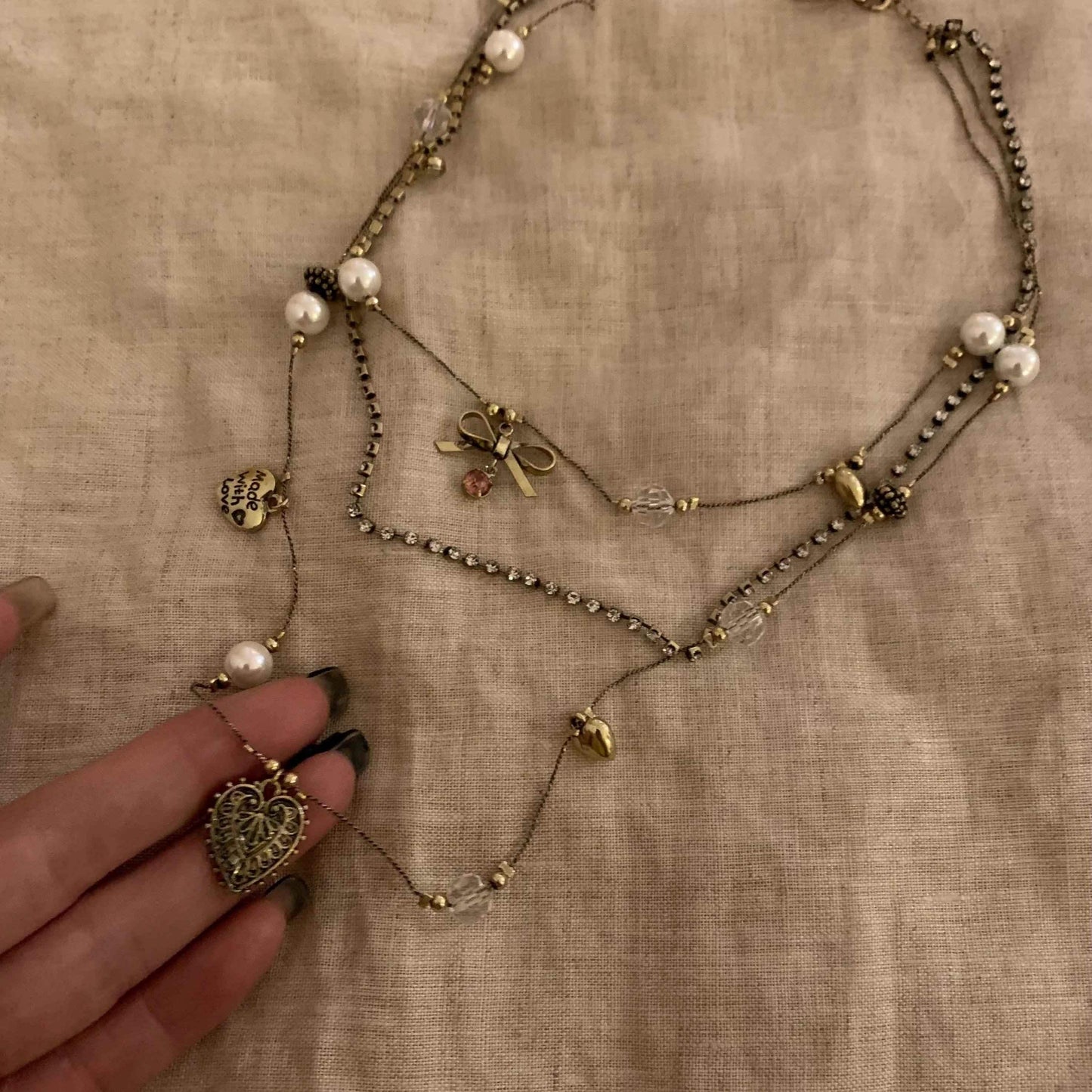 Layered gold necklace with pearls, heart charms, and bow pendant on linen background