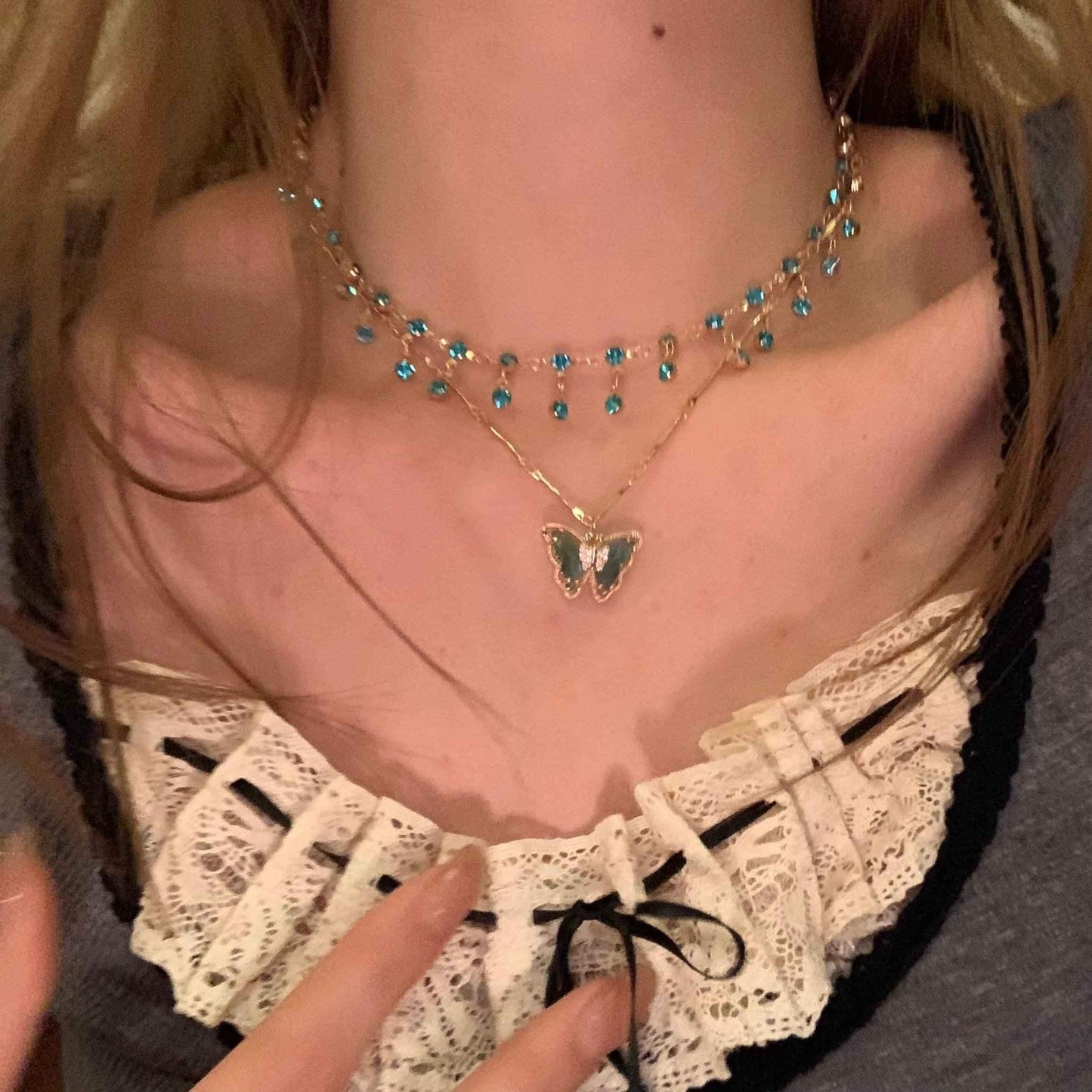 Layered gold necklace with blue crystal choker and green butterfly pendant, worn over lace top
