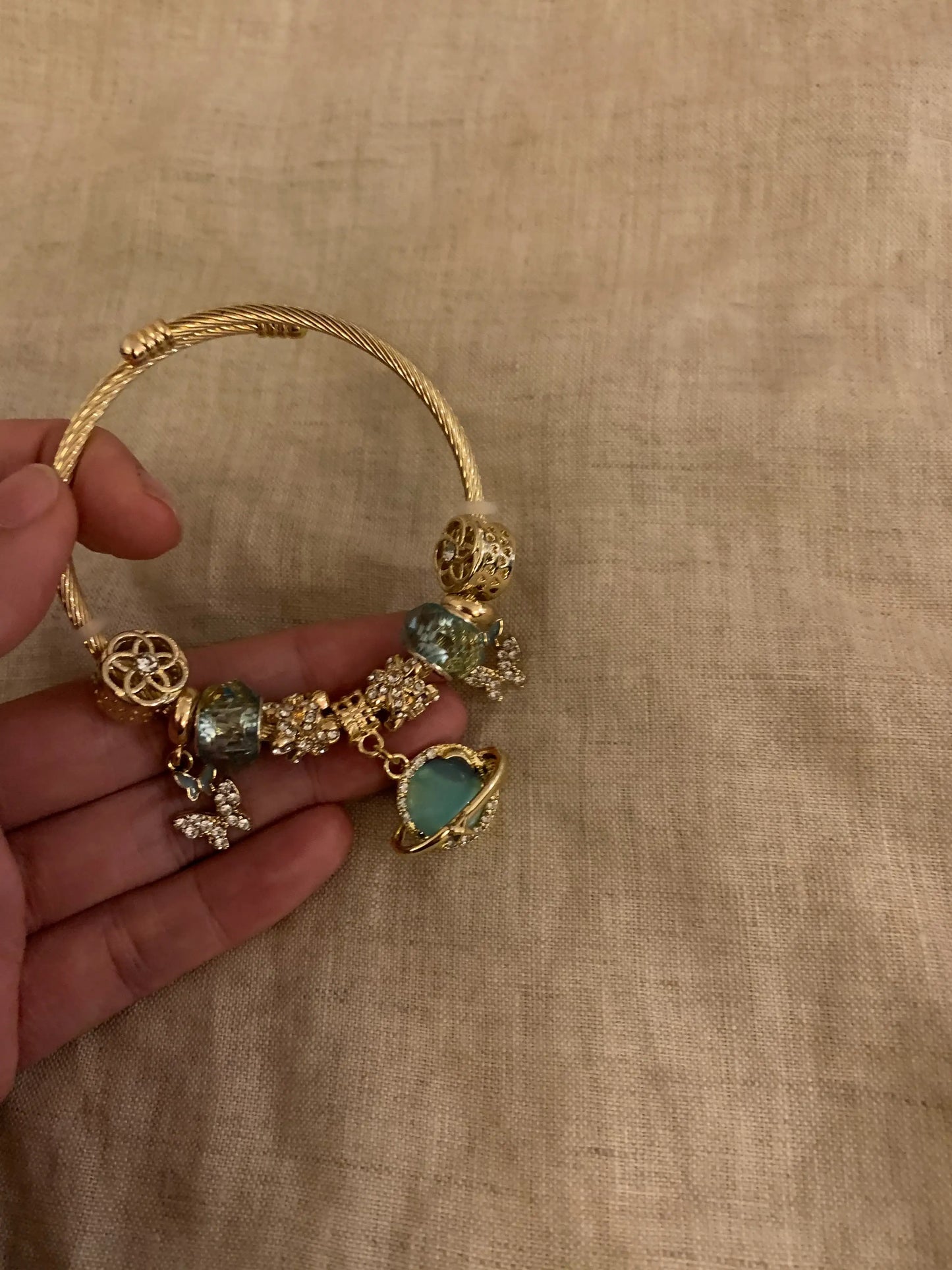 Gold charm bracelet with teal stones and intricate designs, held in hand against fabric background