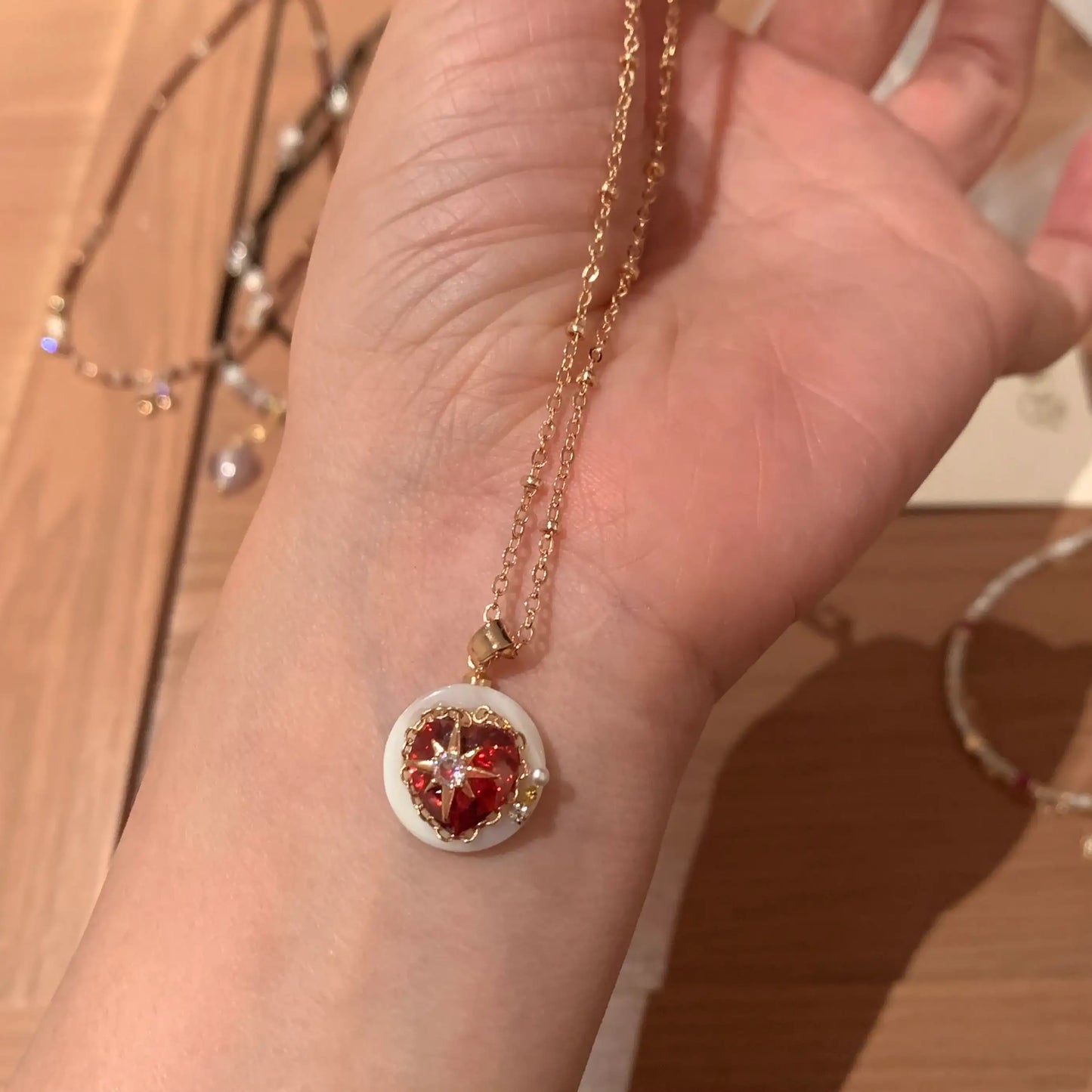 Gold chain necklace with red heart pendant, white circular frame, and delicate star design, held in hand.