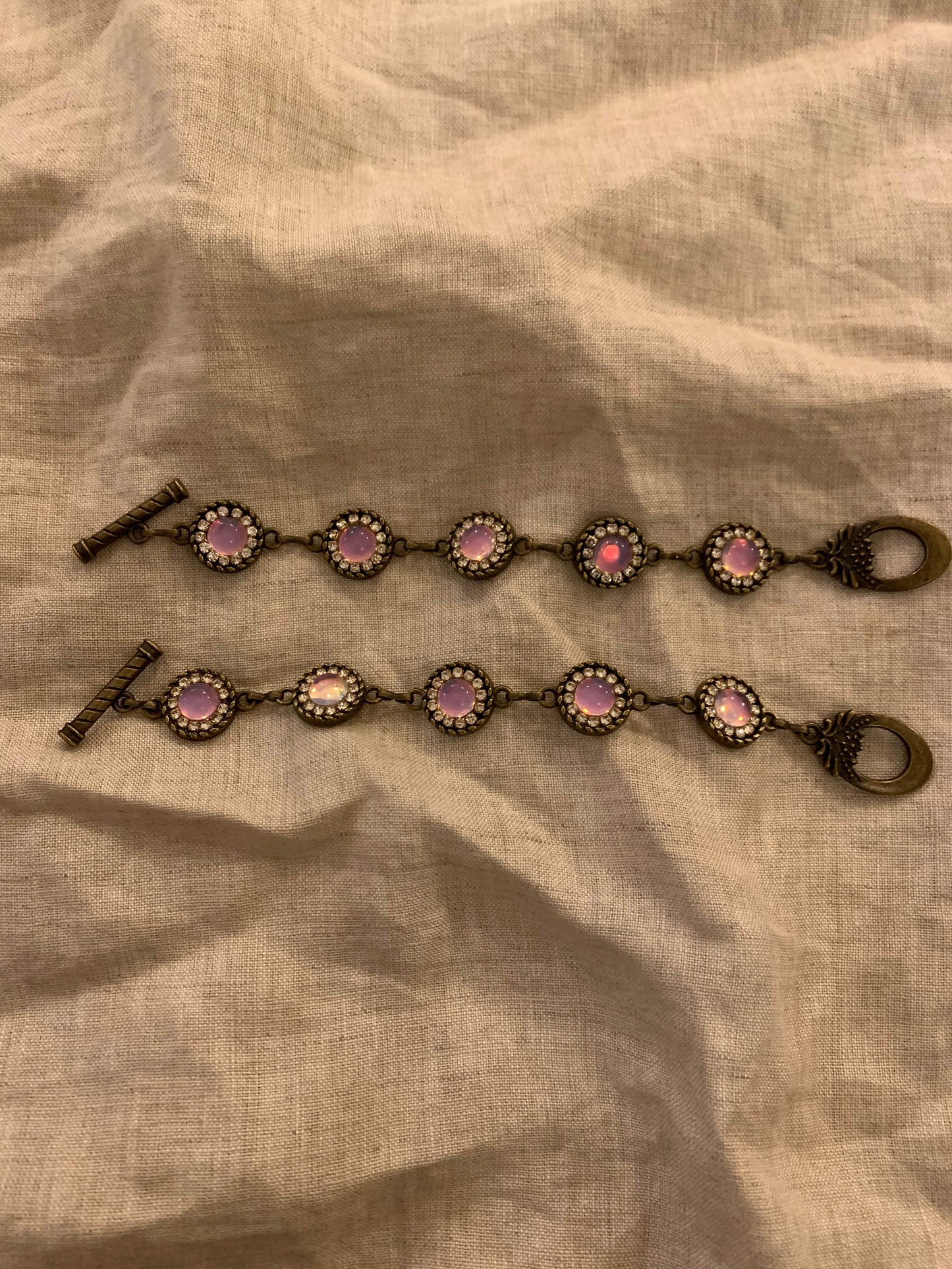 Two vintage-style bracelets with pink opal-like stones and crystal halos on antique brass, laid on linen.