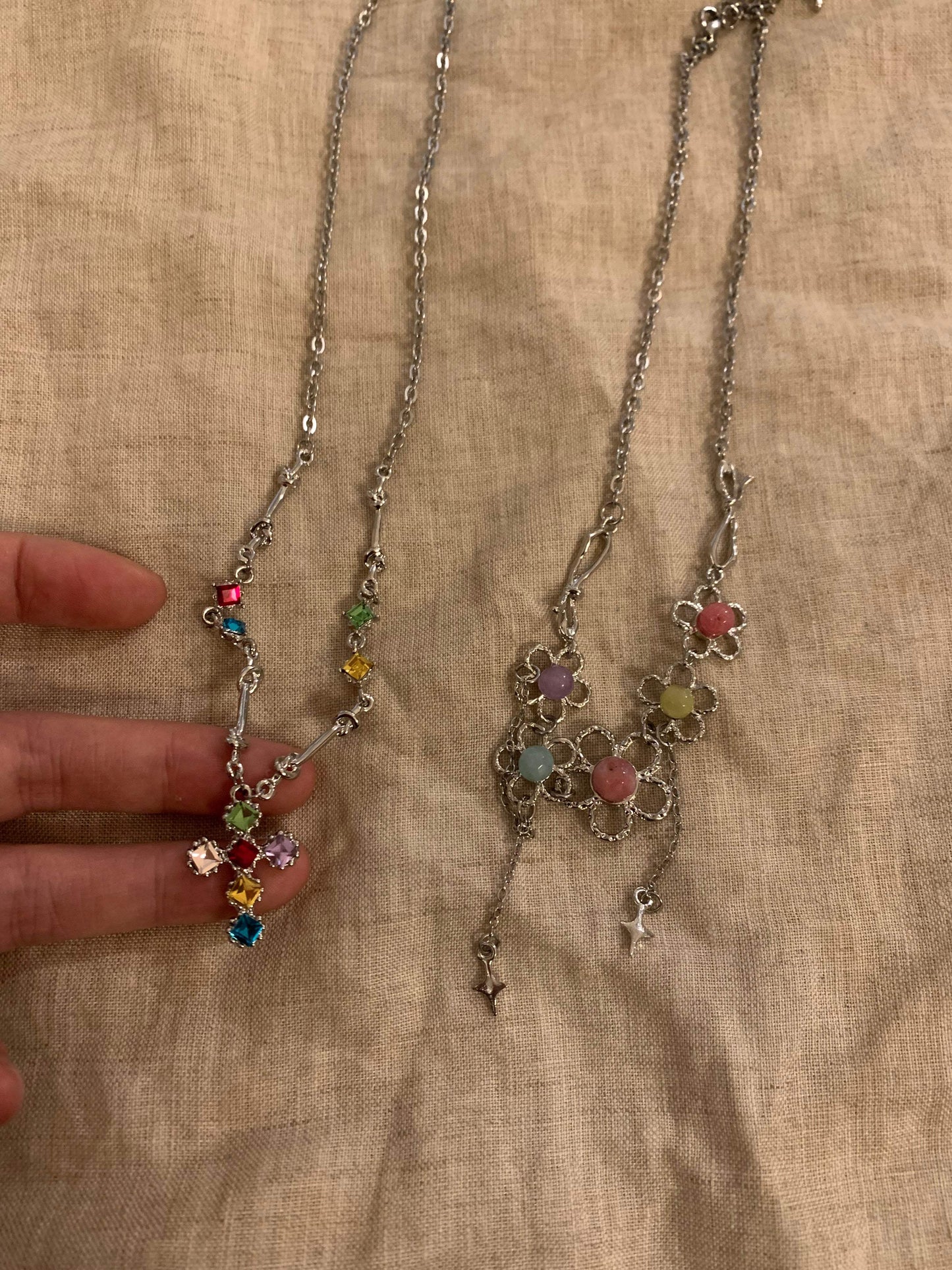 Two elegant necklaces: one with colorful gemstone cross pendant, another with floral design and star charms, displayed on fabric.