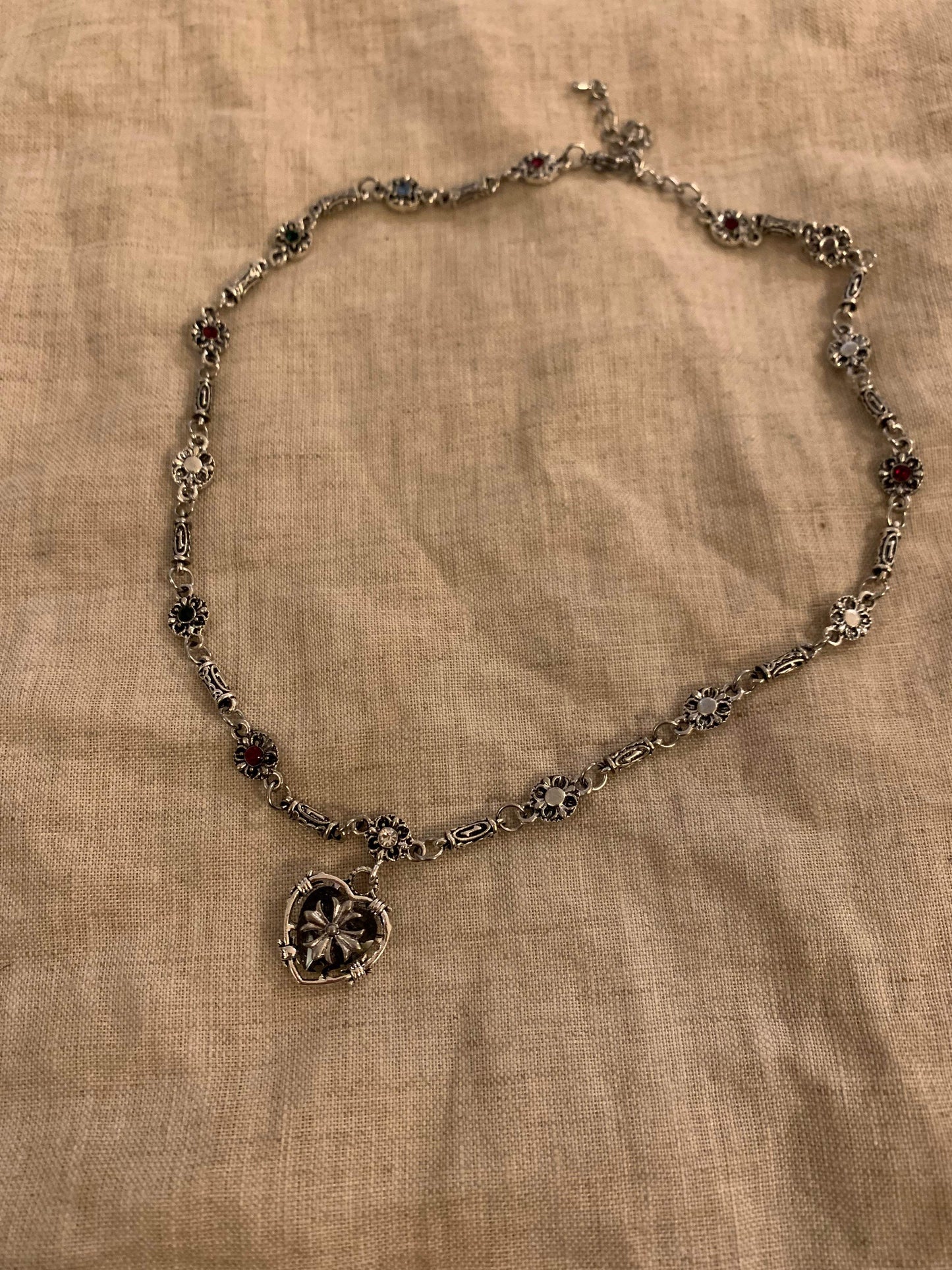Elegant silver necklace with floral links and heart pendant, featuring colorful gemstones on a textured fabric background.
