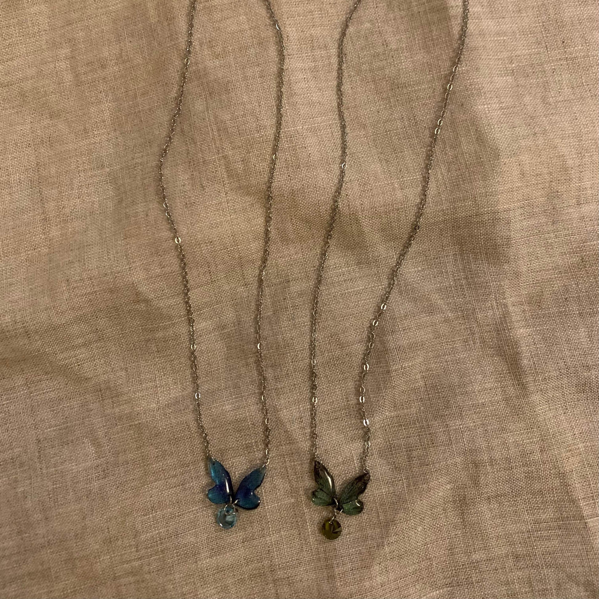 Two butterfly pendant necklaces with blue and green enamel, delicate chains on fabric background