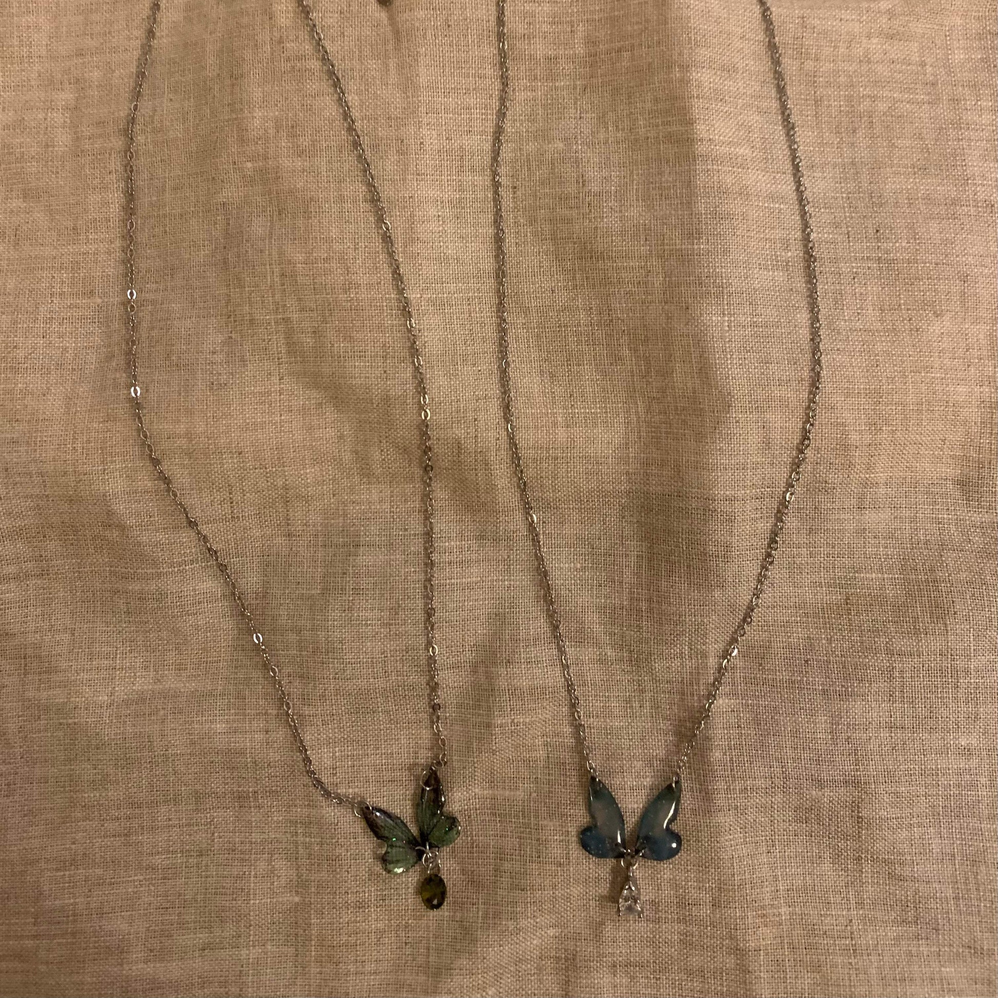 Two delicate butterfly pendant necklaces with green and blue gemstones on a textured fabric background